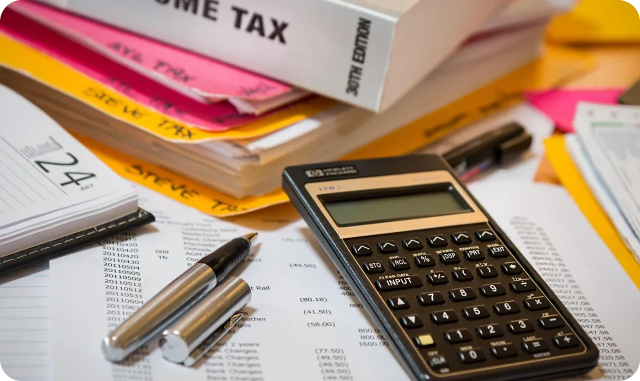 Financial documents, calculator, and pens on a desk with income tax files, representing accounting, bookkeeping, and tax preparation work.