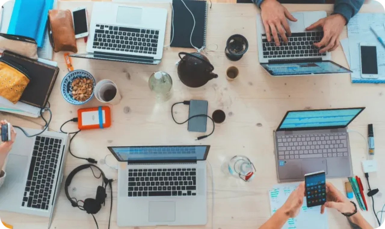 A desk with multiple laptops and accessories and four people working.