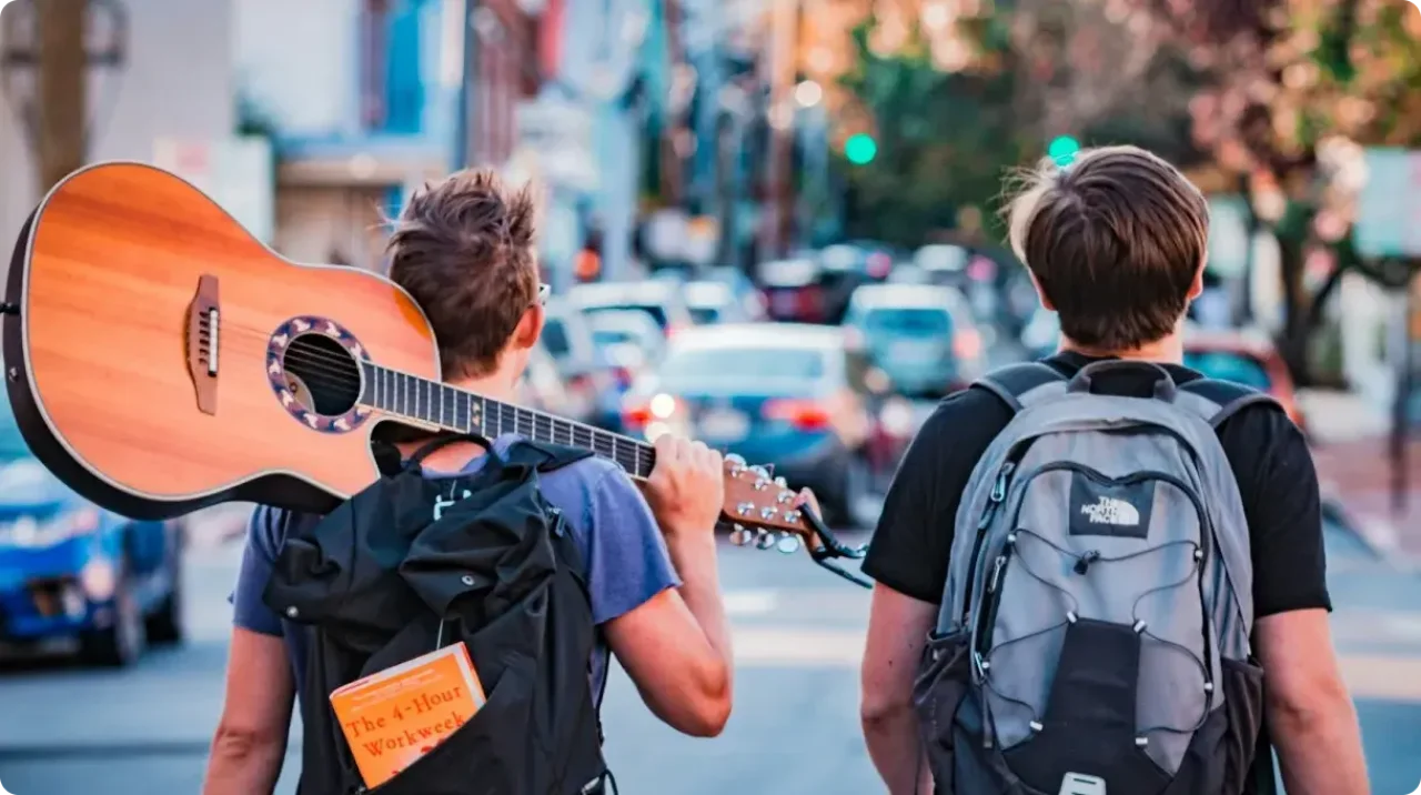 Two Friends Walking In City With Backpacks And Guitar
