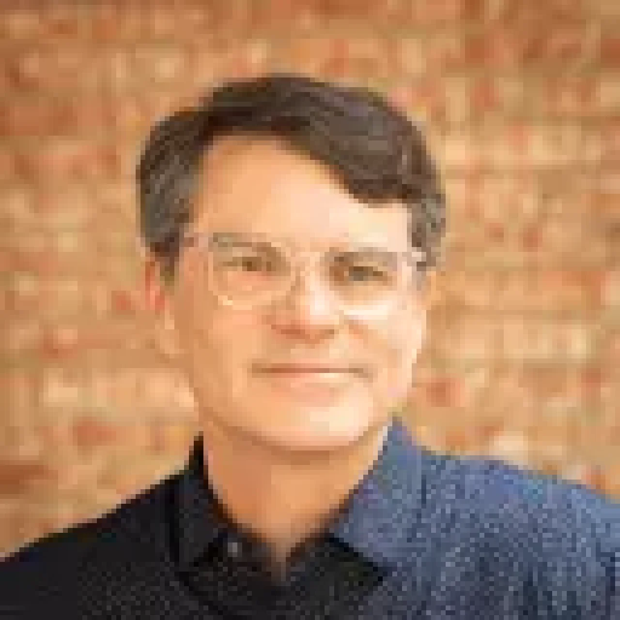 Portrait of middle-aged man wearing glasses and a navy patterned shirt, standing in front of a brick wall background.
