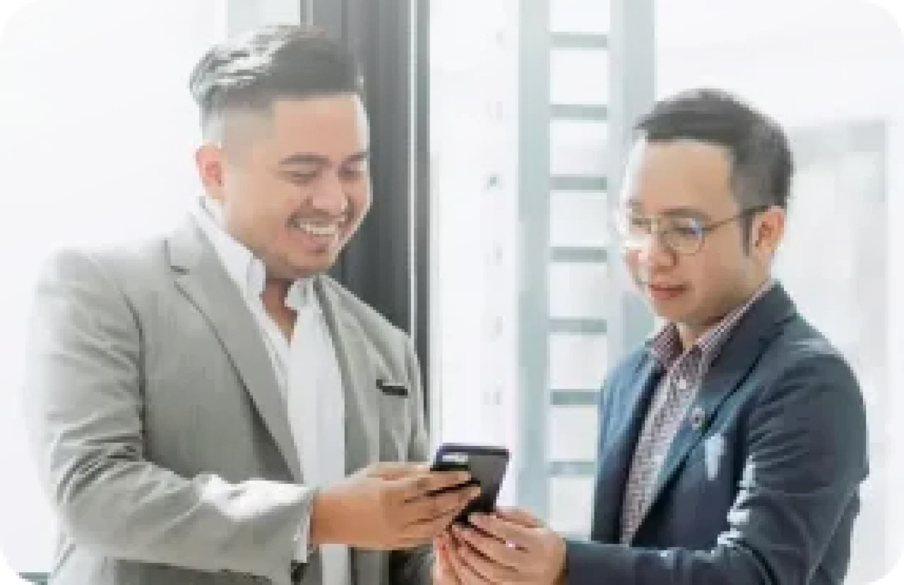 Two businessmen smiling while discussing something on a smartphone next to a bright window
