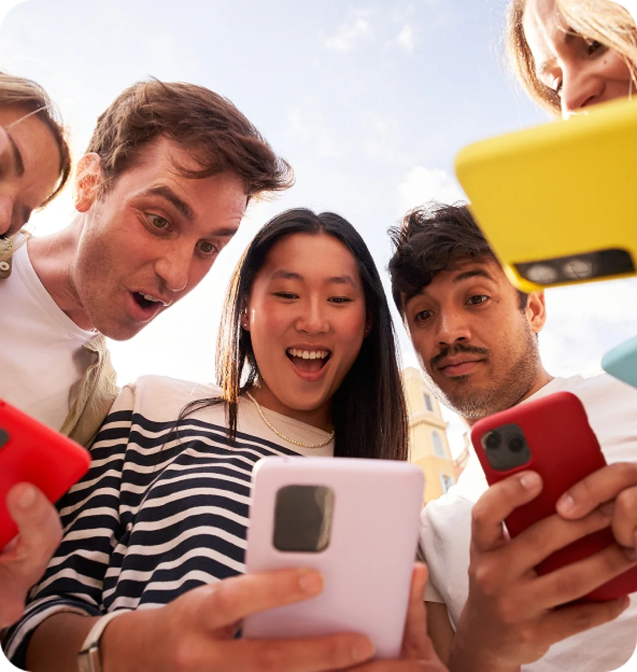 A diverse group of young adults gathered in a circle outdoors, looking excitedly and with surprise at their smartphones, under a bright blue sky.