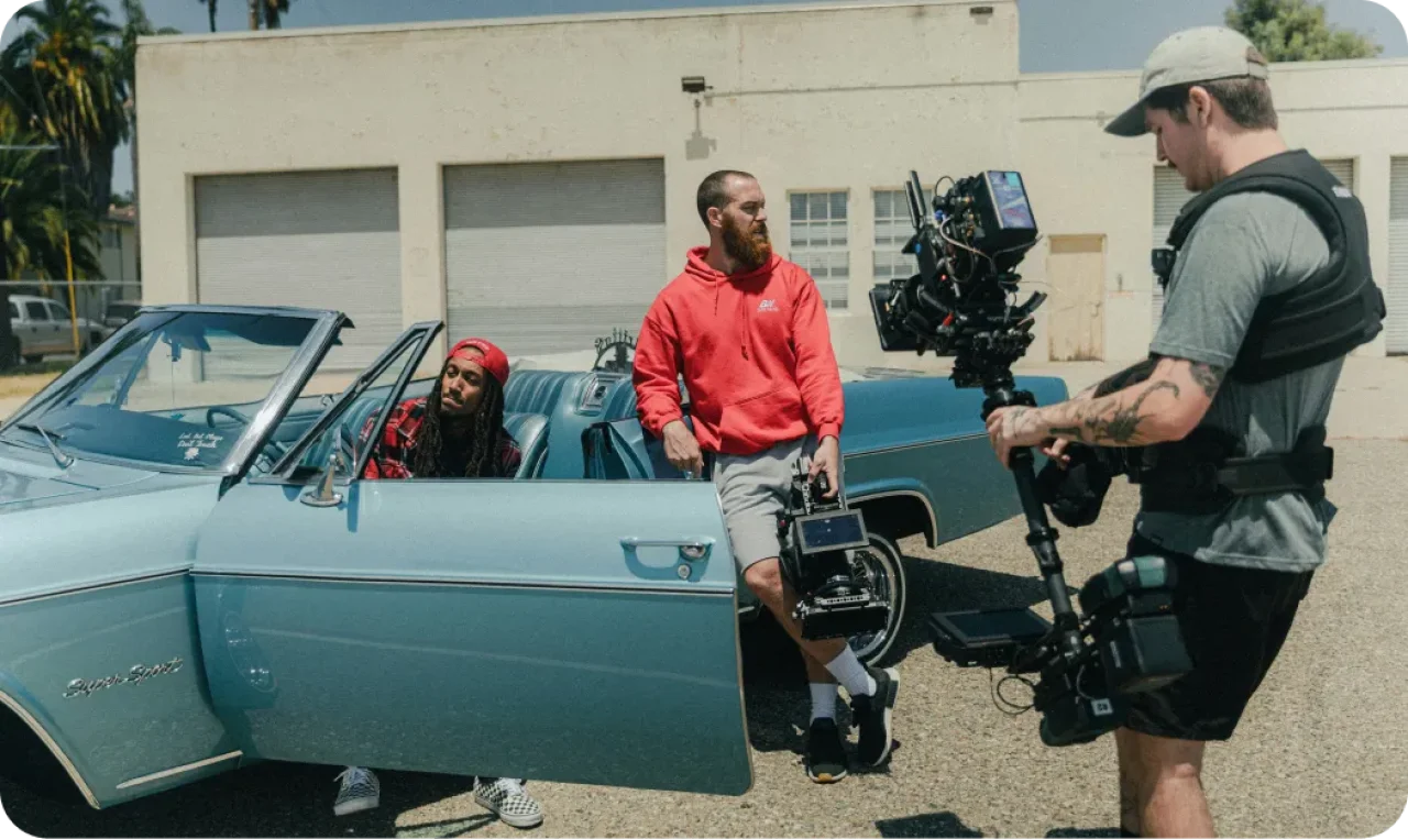 two-men-with-a-classic-blue-convertible-car-while-a-camera-operator-with-a-stabilizer-rigs-a-shot-in-an-outdoor-setting