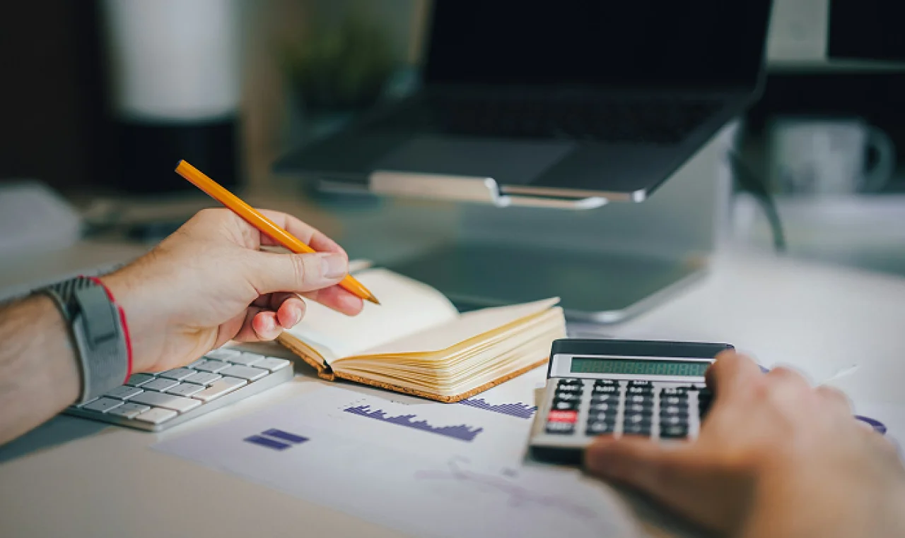 Person working on financial calculations at a desk with a calculator, notebook, and laptop.