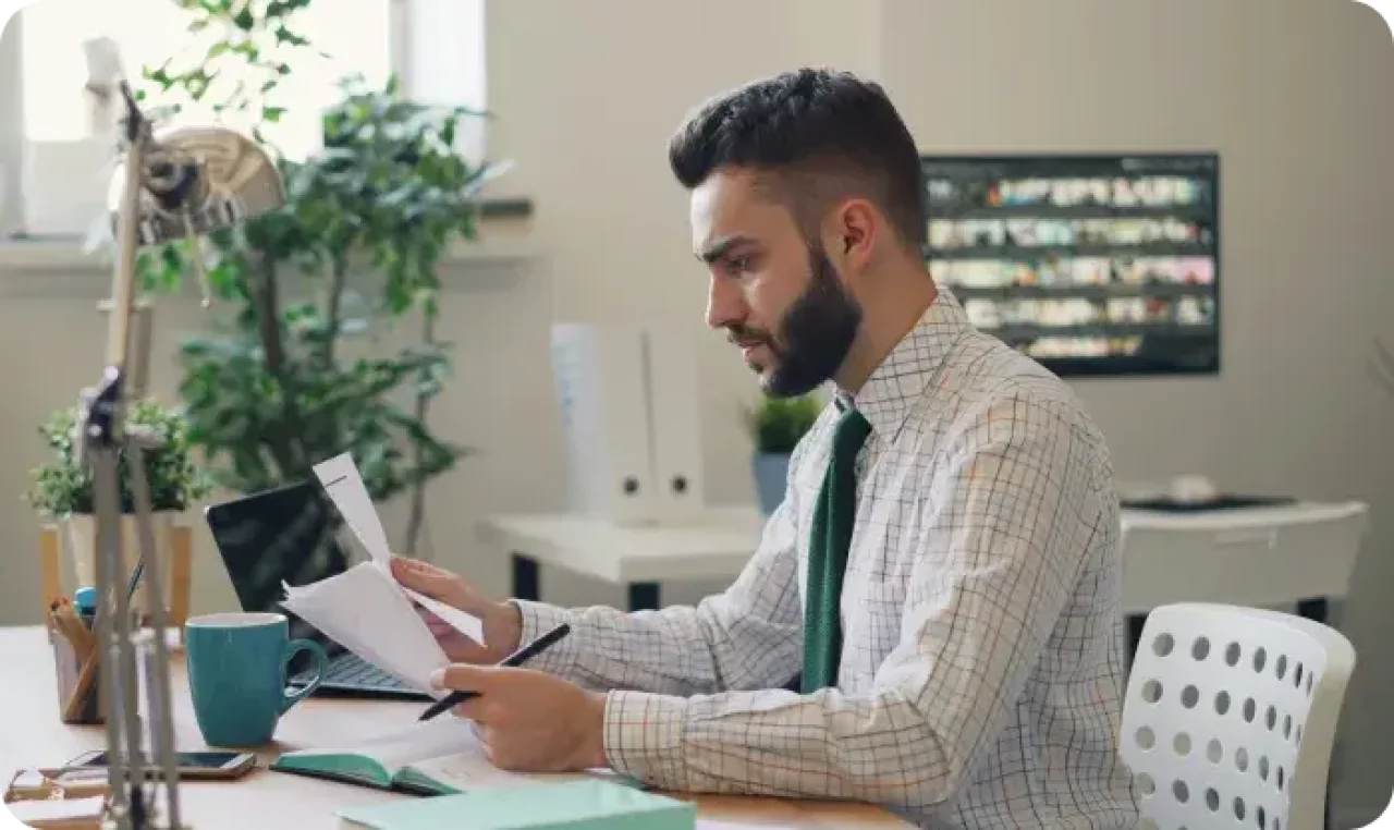 A man in a shirt and tie is seated at a desk, reviewing papers with a focused expression.