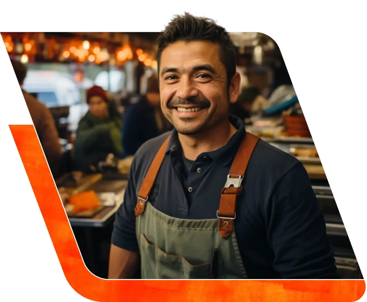 A smiling man wearing an apron stands in a busy market kitchen with warm overhead lights and blurred customers in the background.