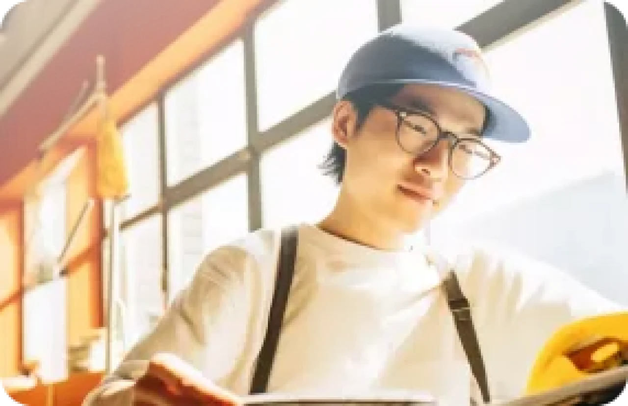 Young student wearing glasses and a blue cap reading a book in a sunlit coffee shop